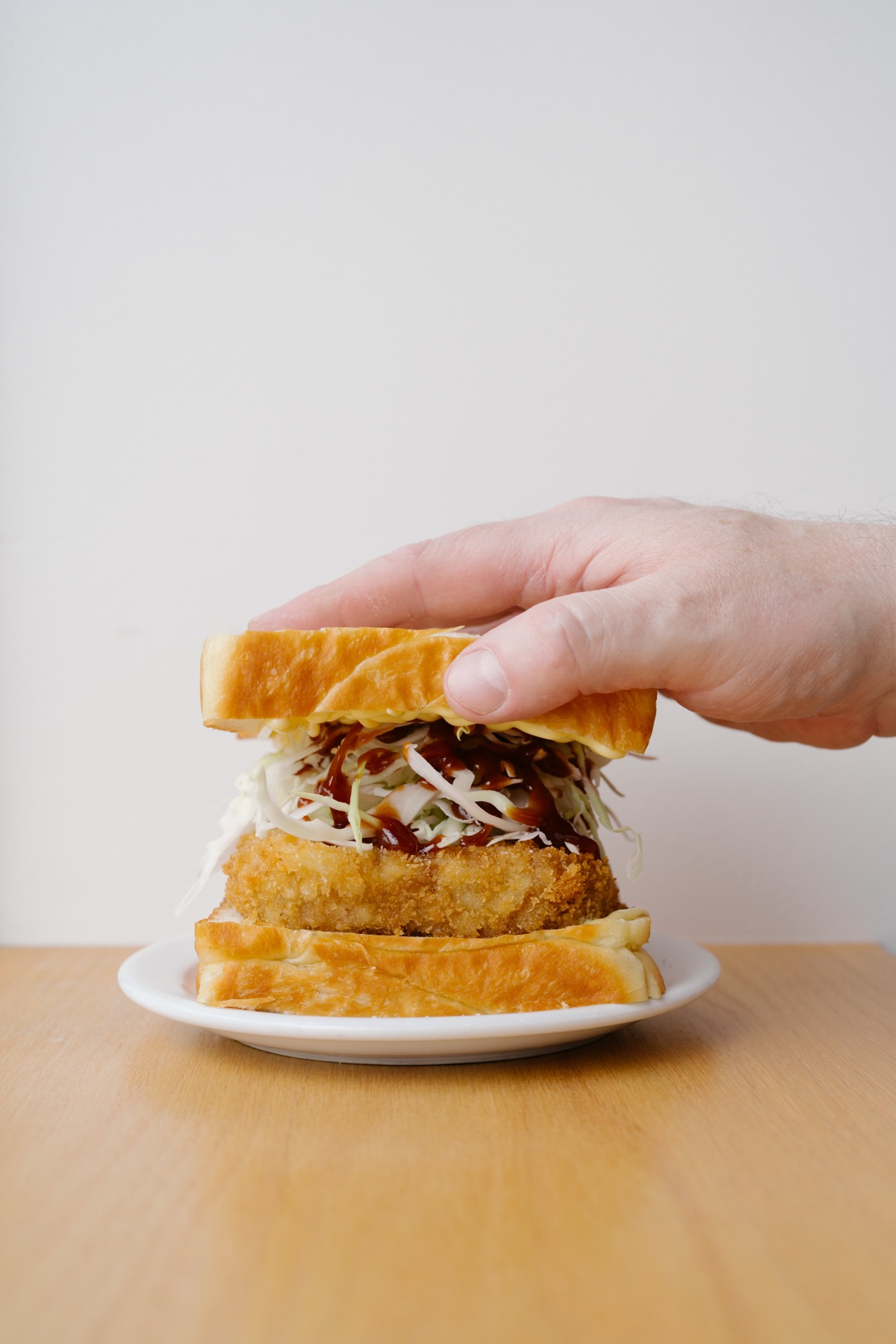 A hand pressing down the top slice of a katsu sando on a white plate